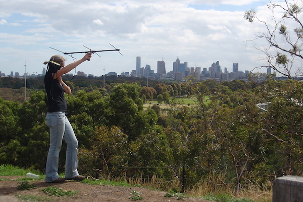 Lisa holding up a radio antenna - behind her is a stretch of parkland, with the Melbourne city skyline in the distance