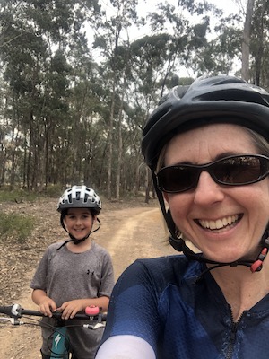 A selfie of Lisa and her son on their bicycles; in the background is a dirt track and trees