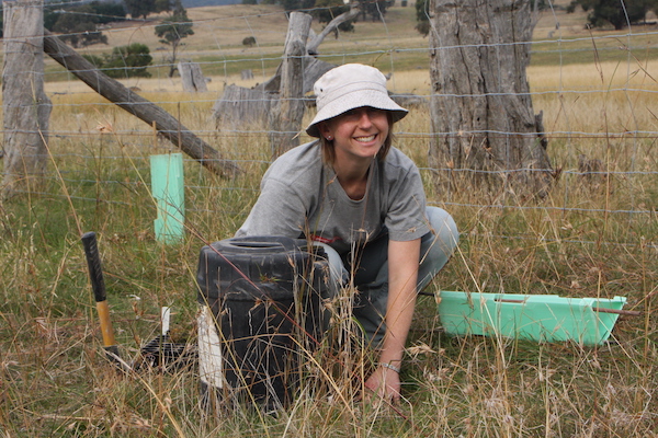 Lisa crouched in long grass with digging equipment and a sample tub beside her; in the background is a barbed-wire fence and rolling paddocks